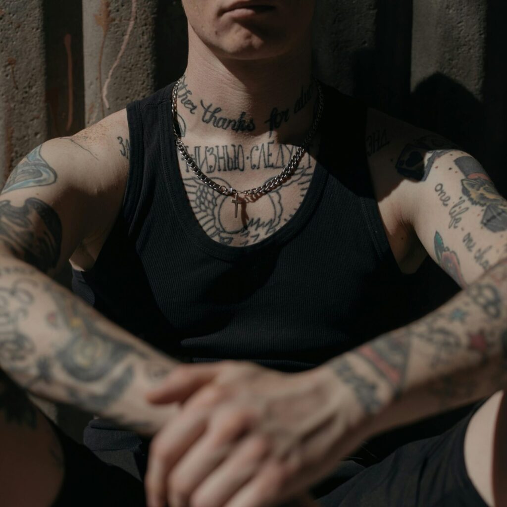 Close-up of a tattooed person in a black tank top sitting against a corrugated wall in moody lighting.